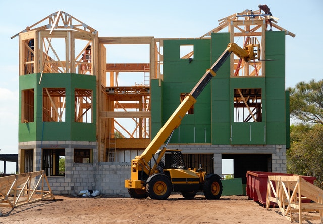 Construction of a custom home in Maryland, featuring a partially built structure with green siding, a construction lift, and workers on the roof.